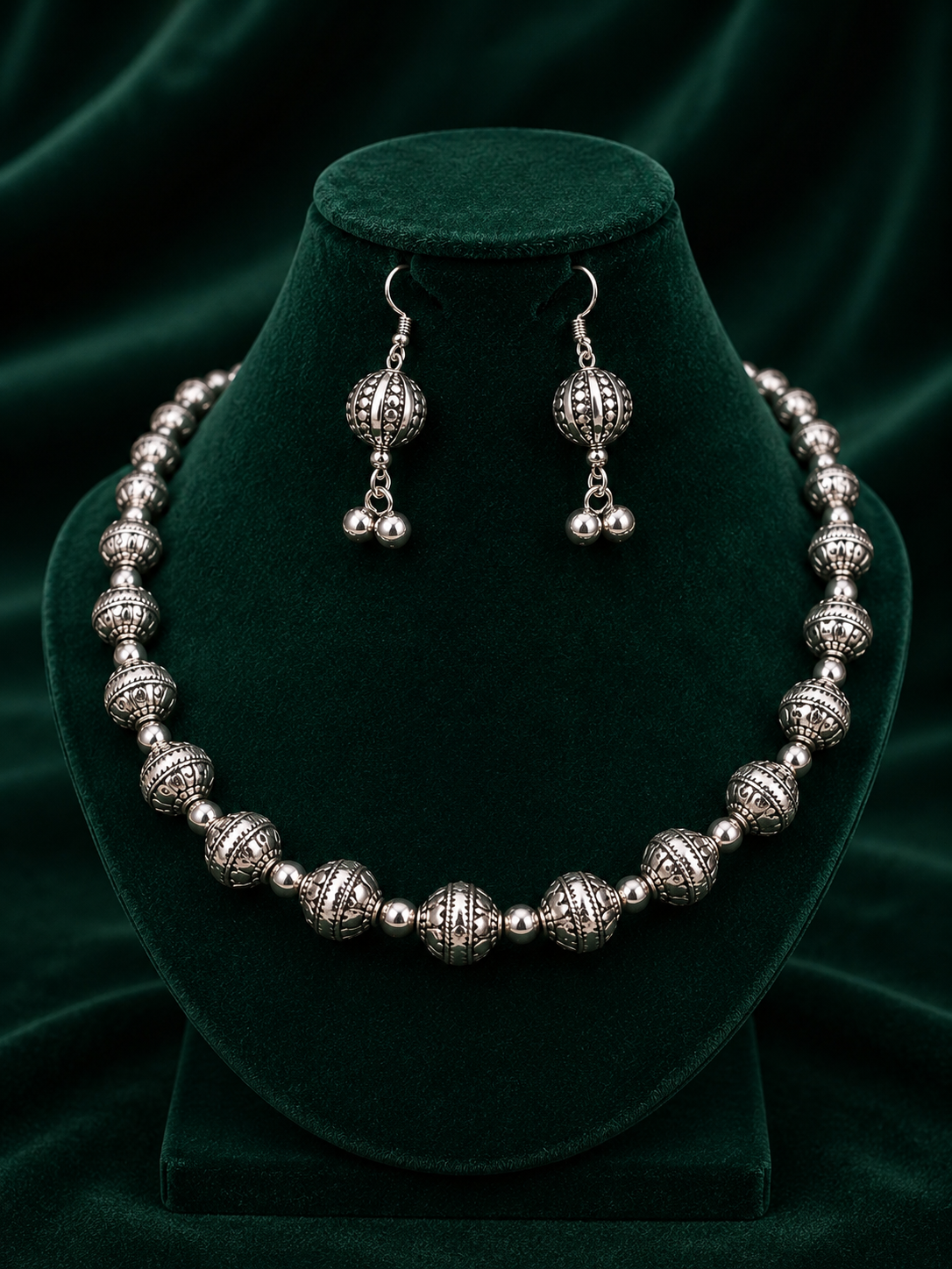 A silver-toned jewelry set featuring a beaded necklace and matching drop earrings, displayed on a dark green velvet jewelry bust against a dark green velvet background.