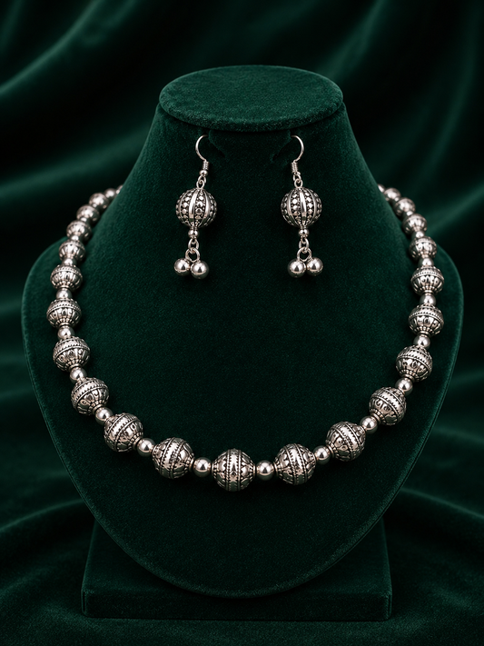 A silver-toned jewelry set featuring a beaded necklace and matching drop earrings, displayed on a dark green velvet jewelry bust against a dark green velvet background.
