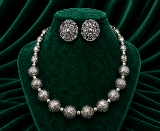 A silver-toned jewelry set featuring a beaded necklace and matching round stud earrings, displayed on a dark green velvet jewelry bust against a draped green velvet background.
