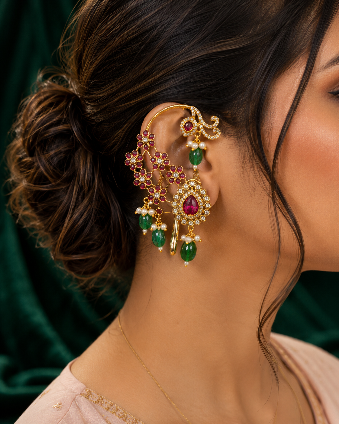 A close-up of a woman's ear wearing an elaborate gold-toned ear cuff and matching dangling earrings adorned with red stones and green teardrop beads, against a dark green velvet background.