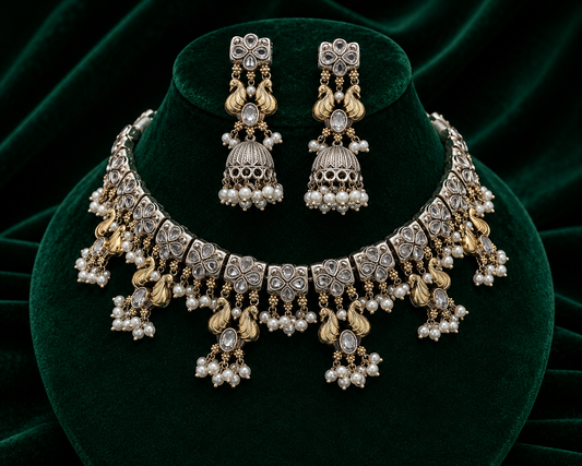 A traditional Indian jewelry set featuring a statement necklace and matching earrings, displayed on a dark green velvet stand. The jewelry is made of silver-toned metal with gold-toned accents, detailed filigree, and clusters of white faux pearls.