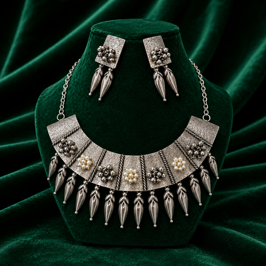 An ornate silver-toned statement necklace with matching drop earrings displayed on a dark green velvet jewelry bust. The necklace features textured silver panels, dangling metallic teardrop charms, and small pearl-like floral accents.
