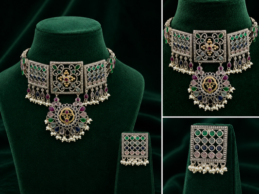An ornate Indian-style jewelry set displayed on dark green velvet mannequin busts, featuring a choker necklace and a matching rectangular pendant. The jewelry is set in silver-toned metal with intricate filigree work, green and purple stones, and small white pearl bead drops.