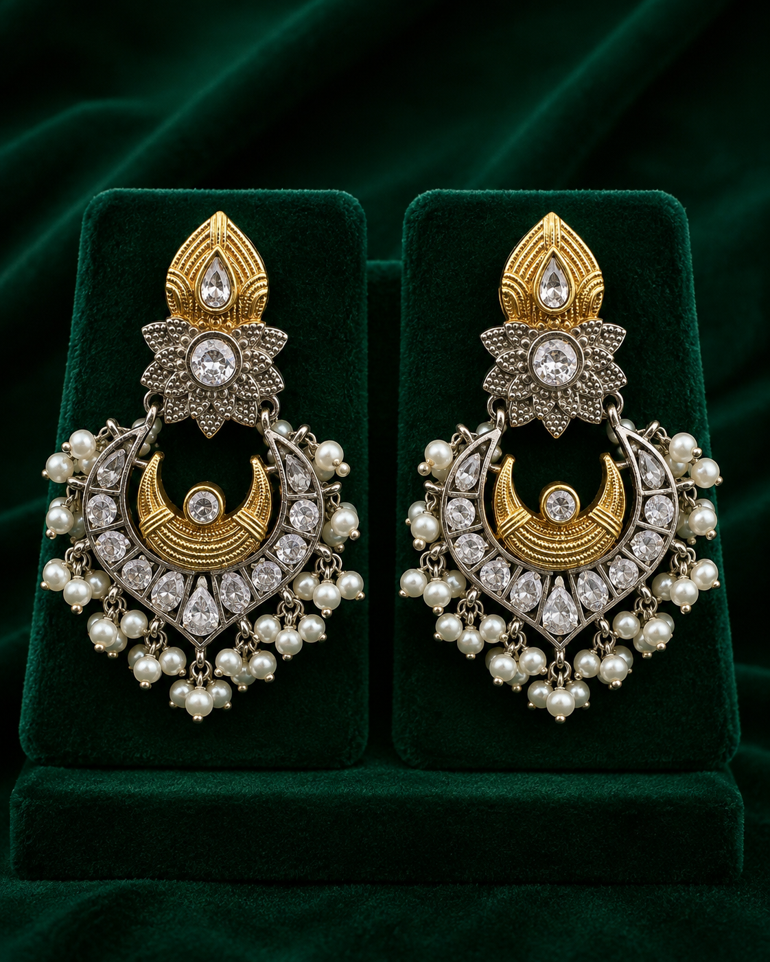 A pair of ornate gold and silver-toned traditional Indian jhumka earrings with white pearls and clear stones, displayed on a dark green velvet jewelry box.