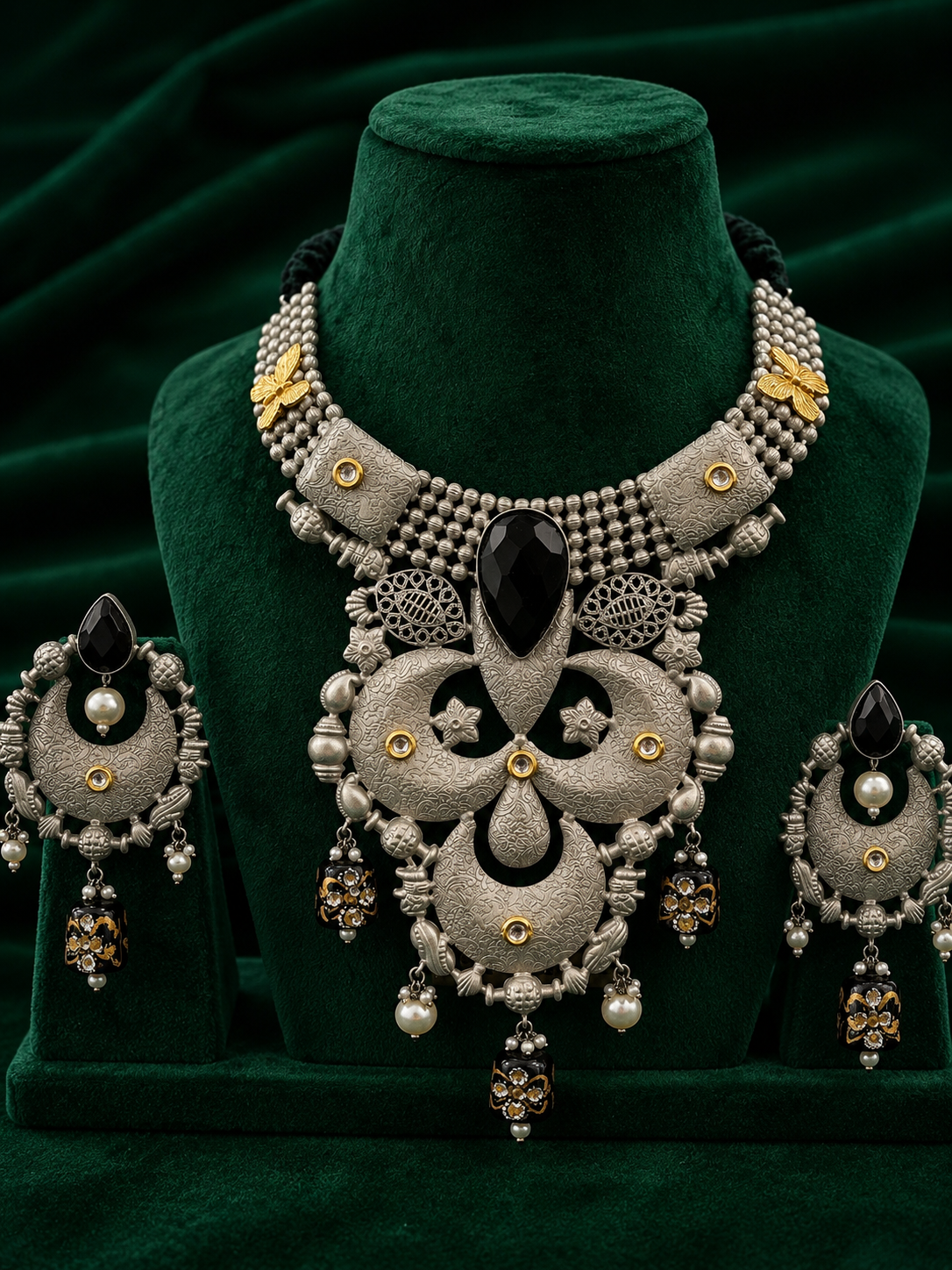 An ornate silver-toned jewelry set displayed on a dark green velvet mannequin bust, including a statement necklace and matching earrings with black teardrop stones, pearl-like white beads, and small gold accents.